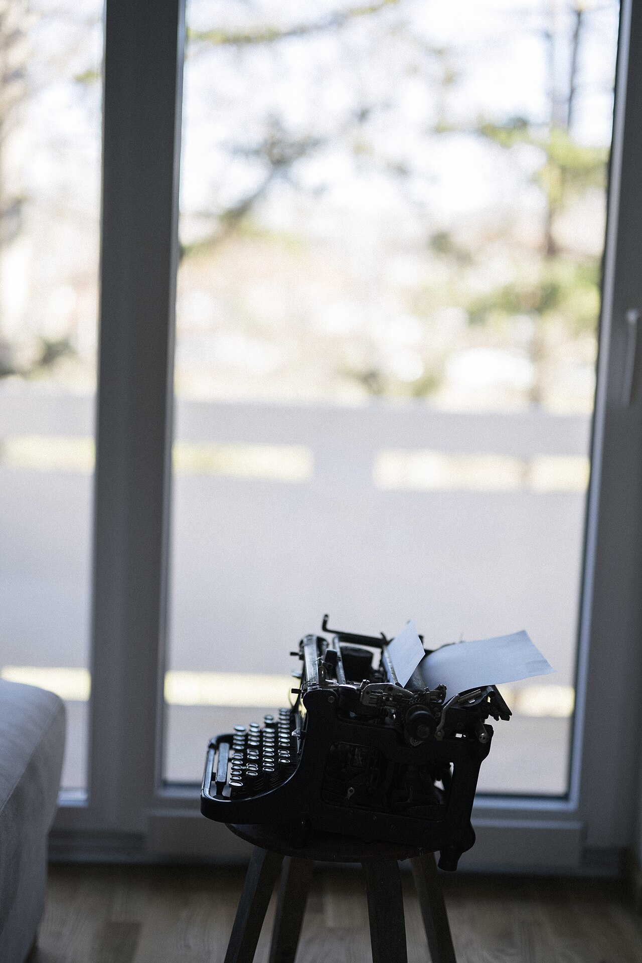 A vintage typewriter resting on a stool beside a large window in a bright, quiet room.